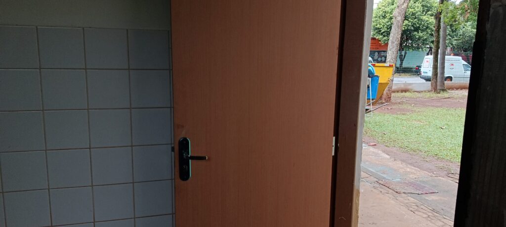 A imagem mostra o interior de um ambiente, possivelmente um banheiro ou cozinha, devido às paredes revestidas com azulejos quadrados de cor clara. À direita, há uma porta de madeira clara entreaberta, com uma maçaneta preta. Através da porta, é possível ver uma área externa com grama, algumas árvores e um caminho de terra. Ao fundo, há uma rua com um veículo branco estacionado e um grande contêiner amarelo. A cena parece ser capturada durante o dia, com luz natural iluminando o ambiente interno e externo. A composição da imagem cria um contraste entre o espaço fechado e o espaço aberto ao ar livre.