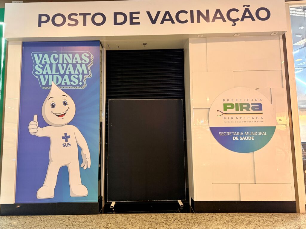 A imagem mostra a fachada de um posto de vacinação localizado dentro de um ambiente que parece ser um shopping ou um centro comercial. A entrada do posto está fechada por uma porta de aço do tipo enrolável, na cor preta. Acima da porta, há uma placa com os dizeres "POSTO DE VACINAÇÃO" em letras grandes e azuis. Ao lado esquerdo da entrada, há um grande cartaz com o personagem Zé Gotinha, mascote tradicional das campanhas de vacinação no Brasil, que está sorrindo e fazendo um gesto de positivo com a mão. No cartaz, está escrito "VACINAS SALVAM VIDAS!" em um balão azul. Do lado direito da entrada, há um logo da Prefeitura de Piracicaba e da Secretaria Municipal de Saúde, com as cores verde e azul, junto do slogan "PIRA PIRACICABA FAZENDO O QUE PRECISA SER FEITO". As paredes ao redor são brancas e possuem um acabamento texturizado. O chão é claro, com um piso que parece ser de pedra ou cerâmica. A iluminação é uniforme e clara, típica de ambientes fechados e comerciais. A imagem transmite a mensagem de um local oficial destinado à vacinação, com destaque para a importância das vacinas para a saúde pública.