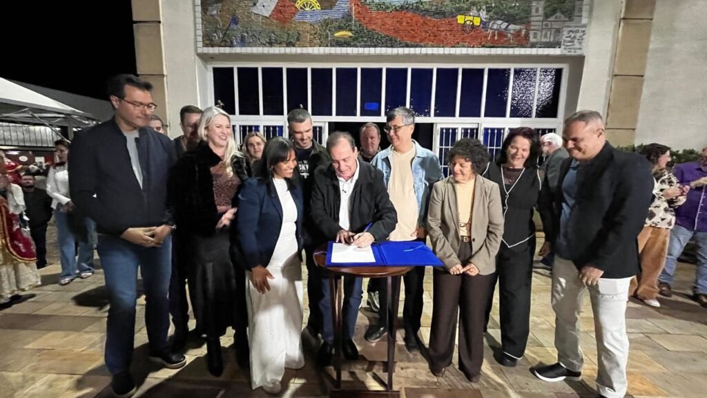 A foto registra um momento de assinatura de documento, aparentemente um ato oficial, durante a noite.Cena principal:Ação central: Um homem de jaqueta preta assina um documento sobre uma pasta azul, apoiada numa mesinha redonda de madeira. Várias pessoas ao redor acompanham e sorriem.Pessoas: Cerca de 10 pessoas estão reunidas em volta da mesa. Homens e mulheres, com roupas sociais e casuais. Destaque para uma mulher de vestido branco longo e blazer azul à esquerda do homem que assina. Todo mundo parece feliz com o momento.Local: O evento acontece ao ar livre, em frente a um prédio com grandes janelas de vidro. Acima das janelas tem um painel de mosaico colorido com desenhos que remetem à história ou cultura local. O chão é de pedra.Atmosfera: É noite e tem mais gente ao fundo, incluindo pessoas com trajes típicos à esquerda. Parece uma solenidade pública, inauguração ou celebração.O contexto sugere a formalização de algum acordo, convênio ou lei, com presença de autoridades e comunidade.