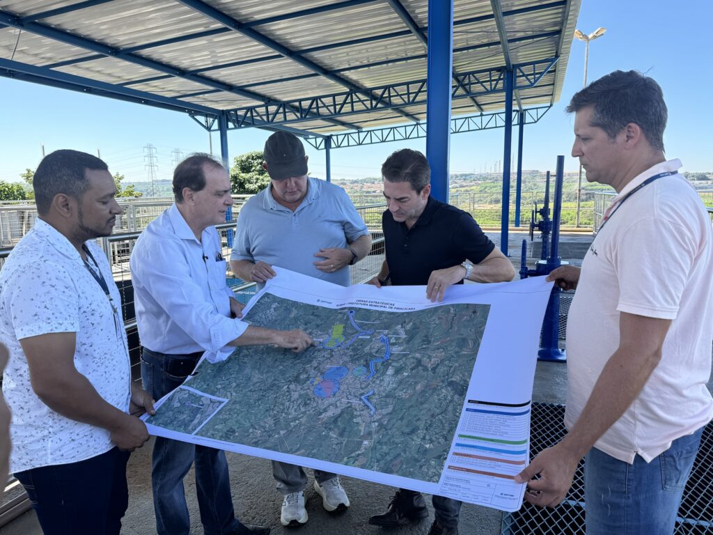 Um grupo de cinco homens está reunido ao ar livre, sob uma estrutura metálica, examinando atentamente um grande mapa ou planta baixa. A imagem, tirada em plena luz do dia, mostra cinco homens adultos engajados em uma discussão sobre um mapa detalhado. O mapa, de grandes dimensões e predominantemente branco, exibe uma representação aérea de uma área geográfica com manchas verdes (vegetação), azuis (corpos d'água ou áreas de projeto), e algumas marcações coloridas em roxo e amarelo. Linhas azuis indicam rotas ou infraestruturas. Na parte inferior direita do mapa, há um bloco de texto visível com títulos como "OBRAS ESTRATÉGICAS" e "SEMAE / PREFEITURA MUNICIPAL DE PIRACICABA", indicando que se trata de projetos municipais. O logotipo da "Semae" também é visível. Os homens estão posicionados ao redor do mapa, que é sustentado por três deles. Da esquerda para a direita: O primeiro homem, à esquerda, tem pele morena e cabelo escuro, curto e bem aparado. Veste uma camisa social de manga curta, branca, com um padrão discreto de pequenas manchas escuras, e calças escuras. Um cordão azul com um crachá pendurado no pescoço. Ele olha fixamente para o mapa, com uma expressão séria. O segundo homem, ao seu lado, tem pele clara e cabelo castanho com entradas. Veste uma camisa social de manga comprida azul clara e calças jeans escuras. Um objeto semelhante a uma caneta ou stylus está no bolso de sua camisa. Ele aponta para uma área específica do mapa com o dedo indicador da mão direita, gesticulando enquanto fala. Sua expressão é focada e concentrada. O terceiro homem, no centro da parte superior do grupo, usa um boné azul escuro ou cinza e uma camisa polo azul clara. Ele tem pele clara e um porte físico robusto. Suas mãos estão posicionadas sobre a barriga, e ele inclina a cabeça para baixo, absorvido na leitura do mapa. No pulso esquerdo, ele usa um relógio escuro. O quarto homem, um pouco à direita do centro, tem cabelo escuro penteado para trás e pele clara. Veste uma camisa polo preta de manga curta e calças escuras. Um relógio de pulseira metálica prateada adorna seu pulso esquerdo e um anel dourado está em seu dedo anelar esquerdo. Ele segura a borda direita do mapa com uma das mãos e olha para baixo, com uma expressão séria. O quinto homem, à direita, tem cabelo grisalho curto e pele clara. Veste uma camisa polo rosa clara de manga curta e calças jeans. Assim como o primeiro homem, ele usa um cordão azul no pescoço com o logotipo "Semae". Ele segura a parte inferior direita do mapa e tem uma expressão atenta, olhando para a área que está sendo discutida. O cenário é um ambiente externo, provavelmente uma instalação industrial ou de serviços públicos, sob uma estrutura de telhado corrugado de metal prateado, sustentado por vigas e pilares de metal azul vibrante. O céu é de um azul claro intenso, sem nuvens visíveis. Ao fundo, pode-se ver uma paisagem com vegetação, linhas de transmissão de energia elétrica (torres com cabos) e casas em uma área urbana distante, sugerindo uma vista panorâmica de uma cidade ou região. À direita, elementos de equipamentos industriais azuis, como tubulações e válvulas, são visíveis, reforçando a ideia de uma instalação de infraestrutura. A luz solar é abundante, criando sombras nítidas e destacando os detalhes. A atmosfera geral da imagem é de profissionalismo, colaboração e foco. Os homens parecem estar em uma reunião de trabalho ou vistoria, discutindo planos e projetos importantes com seriedade e dedicação. A ausência de sorrisos ou gestos casuais reforça o caráter sério do encontro.