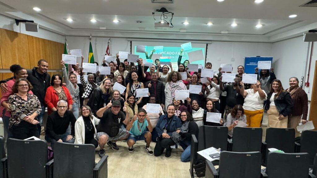 A imagem mostra um grupo grande e diverso de pessoas reunidas em uma sala de conferências ou auditório, todas segurando certificados, indicando que participaram de algum tipo de curso ou evento de capacitação. Elas estão organizadas em várias fileiras, com pessoas em pé ao fundo e sentadas ou agachadas na frente. A maioria está sorrindo, demonstrando satisfação e orgulho pela conquista. Ao fundo, há uma tela de projeção que exibe o texto "CERTIFICADO FASE 1" e possivelmente "logia das 3 fases" (embora parte do texto esteja cortada ou pouco legível). Também há uma faixa vertical à direita com o logo do SEBRAE, uma instituição brasileira que apoia o empreendedorismo. O ambiente é iluminado por luzes no teto, com paredes claras e algumas cadeiras escuras na frente. Essa cena sugere a conclusão de uma etapa de um programa de formação ou treinamento, provavelmente relacionado ao empreendedorismo, dado o contexto do SEBRAE. Todos parecem estar celebrando o sucesso do grupo.