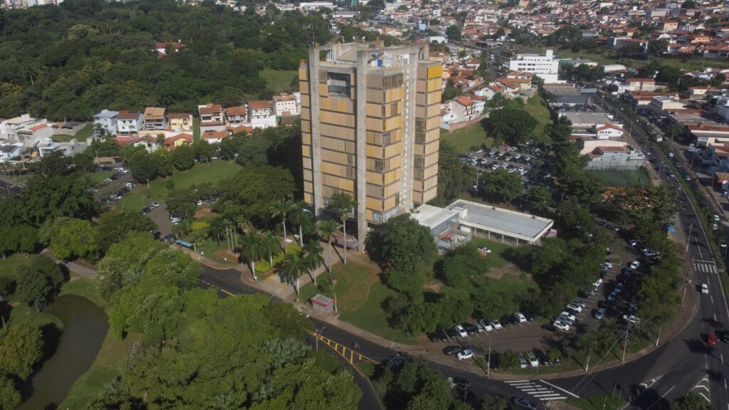 Esta foto aérea mostra um edifício alto e de arquitetura marcante, provavelmente com um estilo brutalista, que se destaca no centro da imagem. Ele tem uma estrutura de concreto aparente e painéis verticais em tons de amarelo e laranja, além de uma base mais ampla. Ao redor do prédio, há bastante área verde com árvores, um estacionamento, ruas com carros circulando e, ao fundo, um bairro residencial com casas e outros prédios menores. O ângulo elevado dá uma boa noção de como esse edifício se integra à paisagem urbana ao seu redor.