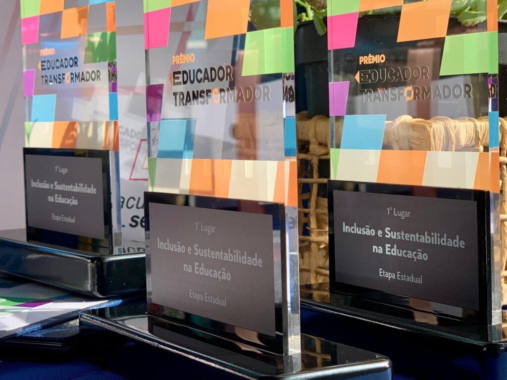 A imagem mostra três troféus de vidro alinhados sobre uma superfície escura. Cada troféu tem uma base preta e uma placa de vidro transparente com detalhes coloridos em forma de quadrados nas partes superior e inferior. No centro do vidro está gravado o texto "PRÊMIO EDUCADOR TRANSFORMADOR". Na base preta de cada troféu, há uma placa com a inscrição "1º Lugar", seguida de "Inclusão e Sustentabilidade na Educação" e "Etapa Estadual". A iluminação destaca o brilho do vidro, dando um aspecto elegante e sofisticado aos troféus. Ao fundo, a imagem está desfocada, sugerindo um ambiente interno, possivelmente um local de premiação ou escritório. Os troféus representam reconhecimento e valorização na área da educação, especificamente em temas de inclusão e sustentabilidade.