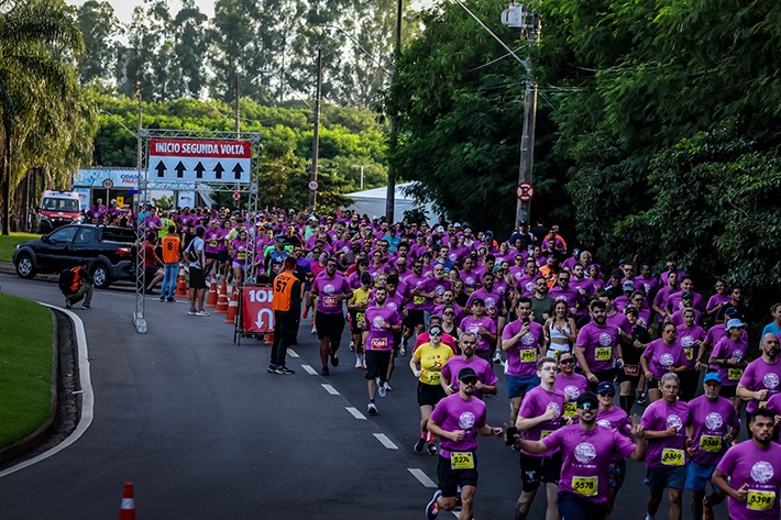 A imagem mostra uma grande quantidade de corredores participando de uma corrida de rua, possivelmente uma maratona ou prova de 10 km. A maioria dos participantes veste camisetas roxas, embora haja algumas pessoas com camisetas amarelas, brancas e outras cores. Eles estão correndo por uma estrada asfaltada, cercada por árvores verdes, o que sugere que a corrida está acontecendo em uma área arborizada ou parque. Na parte da esquerda da imagem, há uma estrutura metálica com uma placa que indica "INICIO SEGUNDA VOLTA 10K", mostrando que os corredores estão no começo da segunda volta da prova. Também é possível ver um veículo preto estacionado próximo e uma pessoa com colete laranja, provavelmente um organizador ou voluntário do evento. O ambiente está iluminado pela luz natural da manhã, e a foto foi tirada de um ângulo elevado, mostrando a multidão de corredores avançando pela estrada. A atmosfera transmite energia, esforço e a sensação de uma competição ou evento esportivo coletivo.