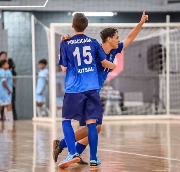 Esta foto mostra dois jogadores de futsal comemorando um gol em uma quadra coberta. • Os jogadores: Eles vestem uniformes azuis idênticos com detalhes brancos. O jogador em primeiro plano está de costas e usa a camisa número 15, com a inscrição "PIRACICABA FUTSAL". O outro jogador está ao lado dele, de frente, com o braço esticado para o alto em gesto de celebração. • O ambiente: Eles estão no meio da quadra, com o piso de madeira típico de ginásios esportivos. Ao fundo, vê-se a rede do gol e outros jogadores do time adversário, também uniformizados, observando a cena. • A atmosfera: É um momento de entusiasmo e alegria típico de uma partida de futsal.
