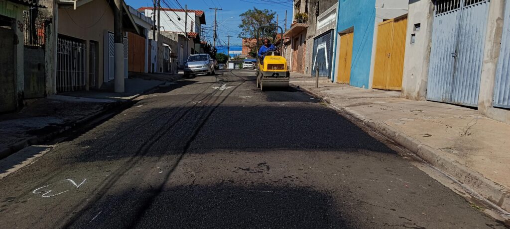 A imagem mostra uma cena urbana durante o dia, onde um rolo compactador amarelo está operando para nivelar e compactar o asfalto recém-aplicado em uma rua. O operador, vestido com uma camisa azul, está sentado no veículo, conduzindo a máquina. A rua é estreita e está cercada por casas residenciais de diferentes estilos e cores, incluindo paredes azuis, bege e de concreto, além de algumas portas de garagem fechadas. Um carro prata está estacionado à esquerda, mais ao fundo na rua. A luz do sol é forte, criando sombras longas no chão. No asfalto, é possível ver algumas marcas feitas com giz, provavelmente para orientar o trabalho. A imagem transmite uma sensação de manutenção urbana, com foco na melhoria da infraestrutura local.