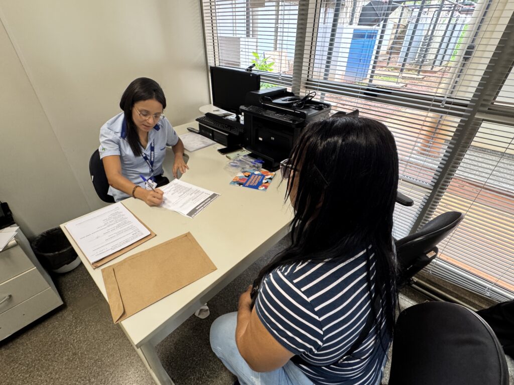Nesta imagem, vemos um atendimento em um escritório. Uma funcionária, usando uma camisa azul clara com um crachá e óculos de grau, está sentada à mesa, preenchendo ou assinando um documento. À sua frente, sentada de costas para a câmera, está uma mulher com cabelos longos e pretos, vestindo uma camiseta de listras horizontais brancas e azuis e calça jeans, aparentemente aguardando o atendimento ou acompanhando o processo. Sobre a mesa, além dos papéis que a funcionária manuseia, há um computador, uma impressora, algumas canetas e um livreto colorido com a inscrição "MIGRANTES, IMIGRANTES E REFUGIADOS". O ambiente é iluminado pela luz natural que entra por uma janela grande com persianas horizontais, através das quais se pode ver uma área externa com portão e grades.
