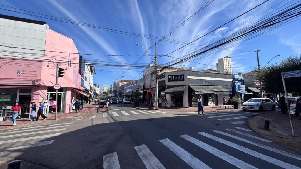 Na imagem, vemos um cruzamento urbano movimentado, com prédios comerciais, pedestres e veículos compondo a cena. À esquerda, destaca-se um edifício rosa com vitrines exibindo manequins e placas de loja, enquanto ao centro aparece um prédio em preto e branco com a fachada da “Jade Ótica”. As ruas possuem faixas de pedestres e semáforos, e várias pessoas caminham pelas calçadas, reforçando o dinamismo do espaço. No alto, fios elétricos e telefônicos cruzam o céu azul com nuvens claras, e à direita uma placa indica a rua “Governador Pedro de Toledo”. O cenário transmite a atmosfera típica de um centro urbano ativo, marcado pela presença de comércio local e pela circulação constante de pedestres.
