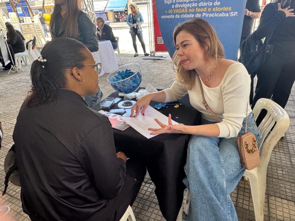 A imagem mostra um cenário ao ar livre, provavelmente em um evento de recrutamento ou feira de empregos. Duas mulheres estão sentadas frente a frente em uma mesa coberta por uma toalha preta. A mulher à direita, loira, veste uma blusa de manga comprida clara e calça jeans azul clara, e está falando, gesticulando com as mãos. A mulher à esquerda, de cabelos curtos e escuros, usa uma blusa preta e óculos, e está ouvindo atentamente. Sobre a mesa há uma tigela transparente com balas embrulhadas em papel azul e alguns papéis. Ao fundo, é possível ver outras pessoas em pé e um banner azul com texto em português que menciona vagas de emprego na cidade de Piracicaba/SP. A cena transmite uma atmosfera profissional e informal, com luz natural do dia.