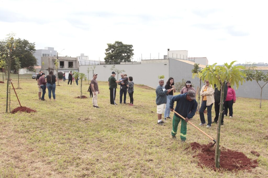Nesta imagem, um grupo de pessoas está reunido em um terreno gramado, participando de uma atividade de plantio de mudas de árvores. O dia está nublado, com luz suave. Em destaque, no canto inferior direito, um homem vestindo moletom azul e calça verde está usando uma enxada para preparar o solo ao redor de uma pequena árvore recém-plantada, que está amarrada a uma estaca de bambu. Ao redor dele, várias outras pessoas estão espalhadas pelo terreno: • Alguns estão em pé, observando o trabalho. • Um homem com uma criança no colo conversa com outras pessoas um pouco mais atrás. • Há um pequeno grupo mais à esquerda, composto por homens e mulheres, que também parecem estar acompanhando ou aguardando sua vez de participar. • Ao fundo, vemos outras mudas de árvores menores já plantadas, marcadas por estacas, e um muro cinza que delimita o terreno, com algumas construções ao longe. O clima parece ser comunitário e colaborativo.