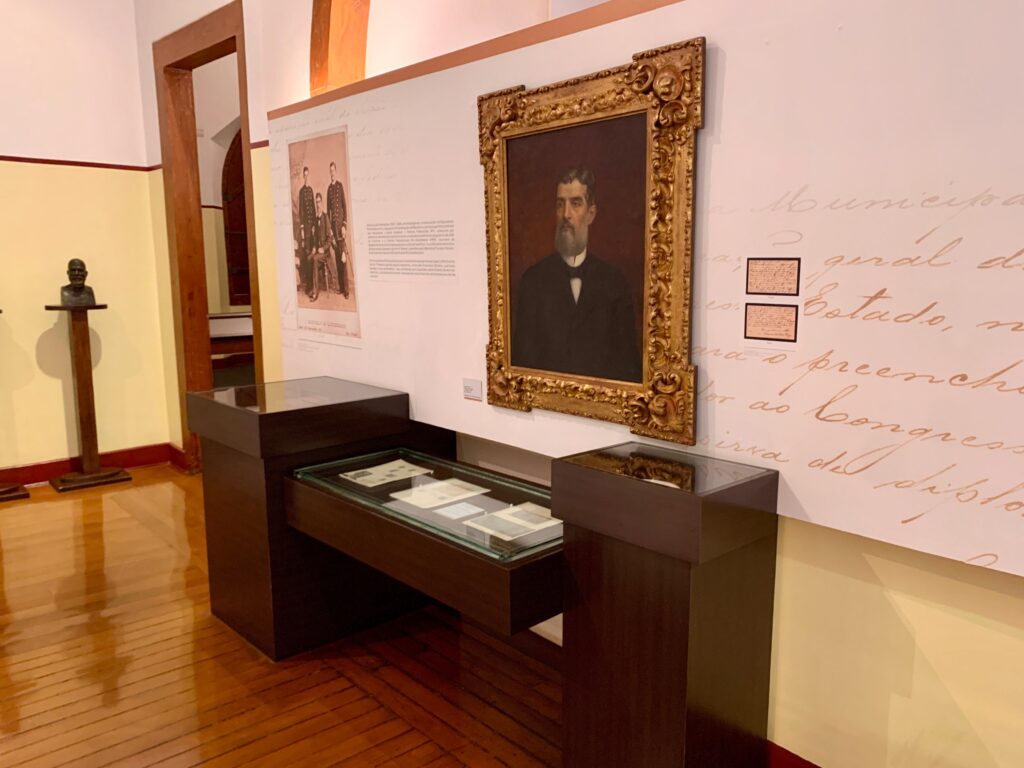 A imagem mostra o interior de uma sala de exposição em um museu histórico, com foco em uma parede decorada com itens relacionados a uma figura política brasileira. * No centro da parede, destaca-se um *retrato a óleo* de um homem de barba, emoldurado em um estilo clássico e ornamentado. * À esquerda do quadro, há uma fotografia antiga em preto e branco que retrata um grupo de homens em trajes formais, acompanhada por um texto explicativo fixado na parede. * Abaixo das imagens, encontra-se uma vitrine de exposição de madeira escura com tampo de vidro, onde estão dispostos documentos históricos. * À direita do retrato, há dois pequenos documentos emoldurados fixados diretamente na parede, sobre um fundo que apresenta uma caligrafia manuscrita ampliada em tom claro. * No canto esquerdo da sala, um busto de bronze está posicionado sobre um pedestal alto e fino, isolado do restante da composição. O piso é de madeira lustrosa e a iluminação é suave, típica de ambientes museológicos.