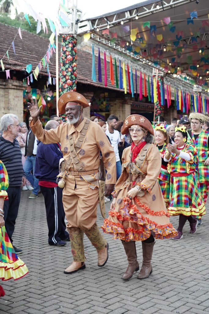 A imagem mostra uma festa típica de São João, conhecida como Festa Junina, que é muito popular no Brasil. No centro da foto, há um casal de idosos vestidos com trajes tradicionais de cangaceiros, que são figuras típicas do Nordeste brasileiro. O homem está acenando com a mão direita e usa um chapéu de couro com detalhes em estrelas, assim como a mulher, que está ao seu lado. Eles vestem roupas marrons com detalhes em couro e costuras decorativas. Ao fundo, outras pessoas também participam da festa, vestindo roupas coloridas e xadrez, típicas desse tipo de comemoração. O local é ao ar livre, com o chão de pedras e uma estrutura metálica decorada com bandeirinhas coloridas penduradas, criando um ambiente festivo e alegre. A iluminação é natural, indicando que a foto foi tirada durante o dia. A cena transmite a cultura popular brasileira e o clima de celebração típico das festas juninas.