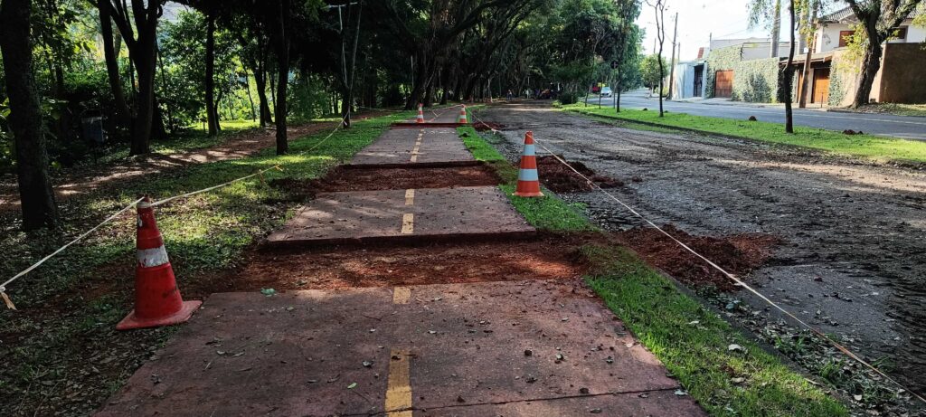 A imagem mostra uma via de pedestres ou ciclovia em processo de reforma ou manutenção, situada ao lado de uma estrada de terra batida e arborizada. * O pavimento avermelhado da pista está sendo interrompido por cortes transversais, onde o solo foi removido, expondo a terra vermelha. * Vários cones de trânsito alaranjados e fitas de sinalização estão posicionados para isolar a área em obras e garantir a segurança. * À direita, vê-se uma rua de terra que margeia uma área residencial com muros cobertos por vegetação e casas ao fundo. * O ambiente é cercado por árvores altas em ambos os lados, criando uma atmosfera arborizada sob uma luz de final de tarde.