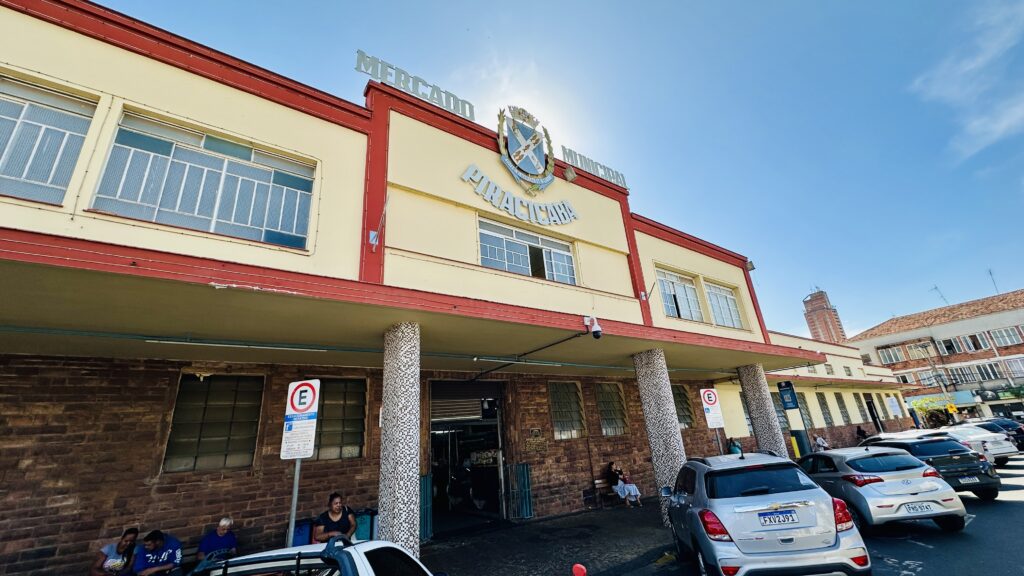 A imagem mostra a fachada do Mercado Municipal de Piracicaba em um dia ensolarado. O edifício tem uma arquitetura clássica, com paredes pintadas de amarelo claro e detalhes em vermelho. Destacam-se as colunas revestidas com pastilhas brancas e pretas que sustentam a parte frontal da estrutura. Na parte superior, há o brasão da cidade e o nome "Mercado Municipal Piracicaba" em destaque. Na parte inferior, vemos uma base de tijolos aparentes, algumas pessoas sentadas próximo à entrada principal e uma fileira de carros estacionados ao longo da rua à direita. O céu está limpo e azul.