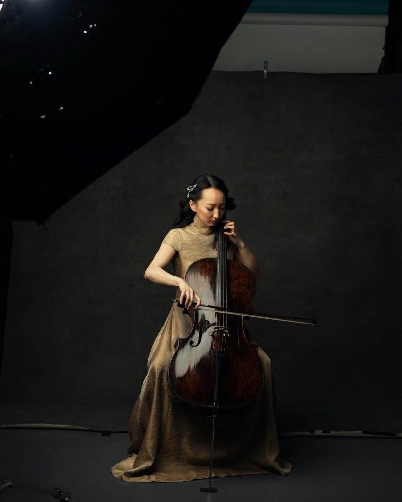 A imagem mostra um retrato profissional em estúdio de uma mulher tocando violoncelo. Ela está sentada, com cabelo escuro preso para trás, vestindo um vestido de gola alta em tom dourado texturizado. A mulher segura o violoncelo de madeira escura e polida, com a mão direita segurando o arco sobre as cordas e a mão esquerda posicionada no braço do instrumento. Seu olhar é focado e sereno. O fundo é neutro e escuro, com textura, e há uma grande softbox visível no canto superior esquerdo, que ilumina a cena com luz suave e direcional. O chão é escuro e fosco. A composição é centralizada e vertical, transmitindo uma atmosfera elegante, artística e sóbria.