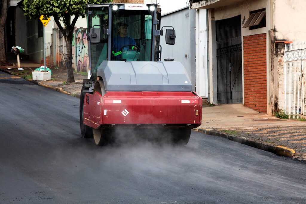 A imagem mostra uma cena de construção ou reparo de uma rua em uma área residencial. No centro da imagem, há uma máquina compactadora de asfalto da marca Dynapac, que está passando sobre uma camada recém aplicada de asfalto quente, de onde sai vapor. O operador da máquina, vestindo colete refletivo amarelo e camiseta azul, está dentro da cabine de vidro da compactadora. Ao fundo, é possível ver uma calçada com árvores, uma placa de sinalização amarela indicando passagem de pedestres, um muro com grafite e algumas residências com portões. A cena parece estar acontecendo durante o dia, com iluminação natural, e transmite a sensação de trabalho em andamento na via pública.