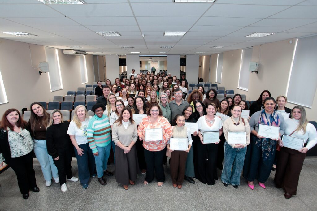 Nesta imagem, um grande grupo de pessoas está reunido em um auditório para uma foto coletiva. A maioria são mulheres, mas há alguns homens também, todos posando em frente às poltronas azuis do local. Muitas das pessoas estão segurando certificados ou diplomas nas mãos, sugerindo que participaram de um curso, treinamento ou cerimônia de conclusão. Elas estão organizadas em fileiras, com as pessoas da frente segurando os documentos e as demais logo atrás, todas sorridentes e olhando para a câmera. O ambiente é um auditório com paredes claras e um piso de granilite, passando uma sensação de um evento formal e organizado.