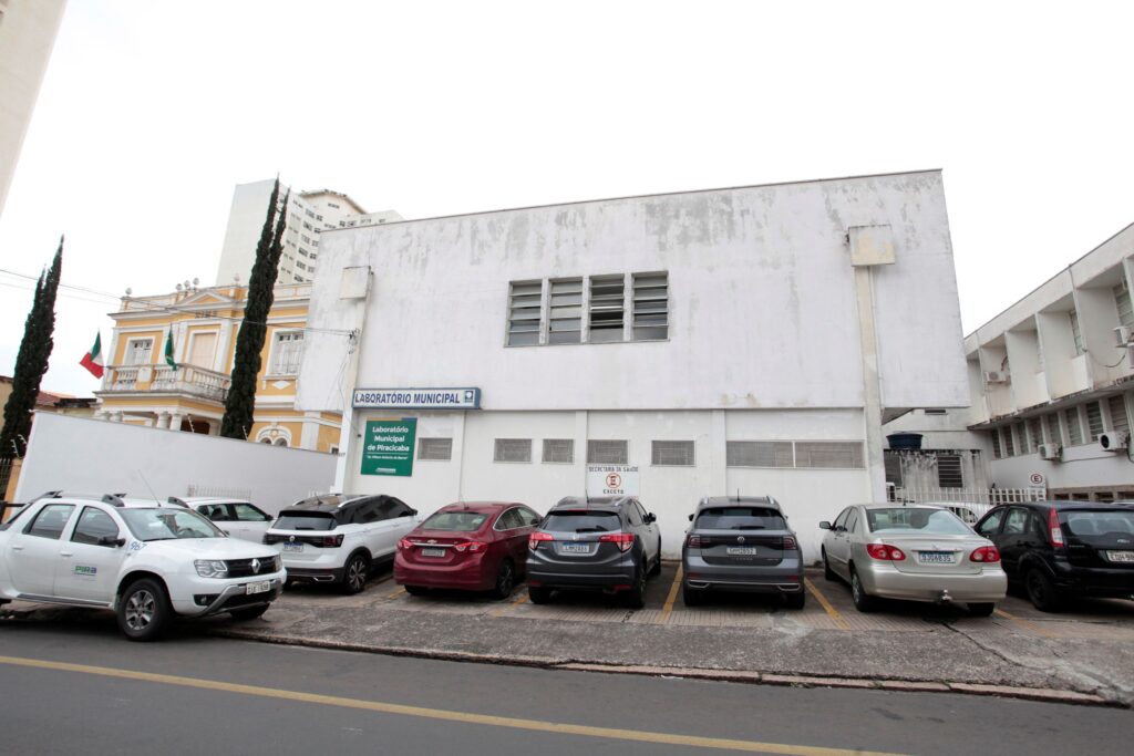 A fotografia externa apresenta a fachada de um laboratório municipal com um estacionamento à frente, revelando um dia nublado com iluminação difusa. A composição da imagem é dominada pela arquitetura do laboratório municipal, um edifício de cor branca com uma textura desgastada, indicando a passagem do tempo. Na frente do edifício, há um estacionamento com diversos veículos estacionados em fila. A ação na imagem é estática, focada na presença dos carros e do prédio. A perspectiva é ligeiramente oblíqua, mostrando tanto a lateral quanto a frente do laboratório. Os carros estão dispostos de forma organizada, preenchendo a maior parte do espaço horizontal da fotografia. Elementos verticais como postes e árvores finas aparecem nas laterais, adicionando um contraste à horizontalidade. Os principais sujeitos são os carros estacionados. Vemos um carro utilitário branco na frente esquerda, seguido por um SUV branco, um sedã vermelho, um SUV preto, um sedã prateado e um hatchback preto à direita. Os carros variam em modelo e cor, mas todos estão em um estado de conservação aparente. A placa de sinalização "Laboratório Municipal" com um símbolo azul e branco é visível na fachada do edifício, indicando a função do local. A imagem é uma fotografia digital com um estilo documental e realista. A técnica de execução foca na captura da realidade sem grandes intervenções artísticas. As cores são contidas, dominadas pelos tons de cinza do céu nublado, o branco do edifício e os diversos tons dos carros. O estilo é direto e informativo, sem embelezamentos, transmitindo a funcionalidade do espaço retratado. A escolha de enquadramento é ampla, abrangendo uma área significativa da rua e do estacionamento em frente ao edifício. O cenário é urbano, com o laboratório municipal como ponto focal. O ambiente ao redor parece ser uma área com outros edifícios, incluindo um prédio histórico com arquitetura amarelada e bandeiras visíveis à esquerda, e um edifício moderno de vários andares ao fundo. A iluminação é difusa e uniforme, característica de um dia nublado, o que resulta em sombras suaves e uma ausência de contrastes fortes. Essa condição de luz cria uma atmosfera neutra e um tanto austera, alinhada à funcionalidade do laboratório. A presença do asfalto na frente e da calçada sugere um ambiente público e acessível.