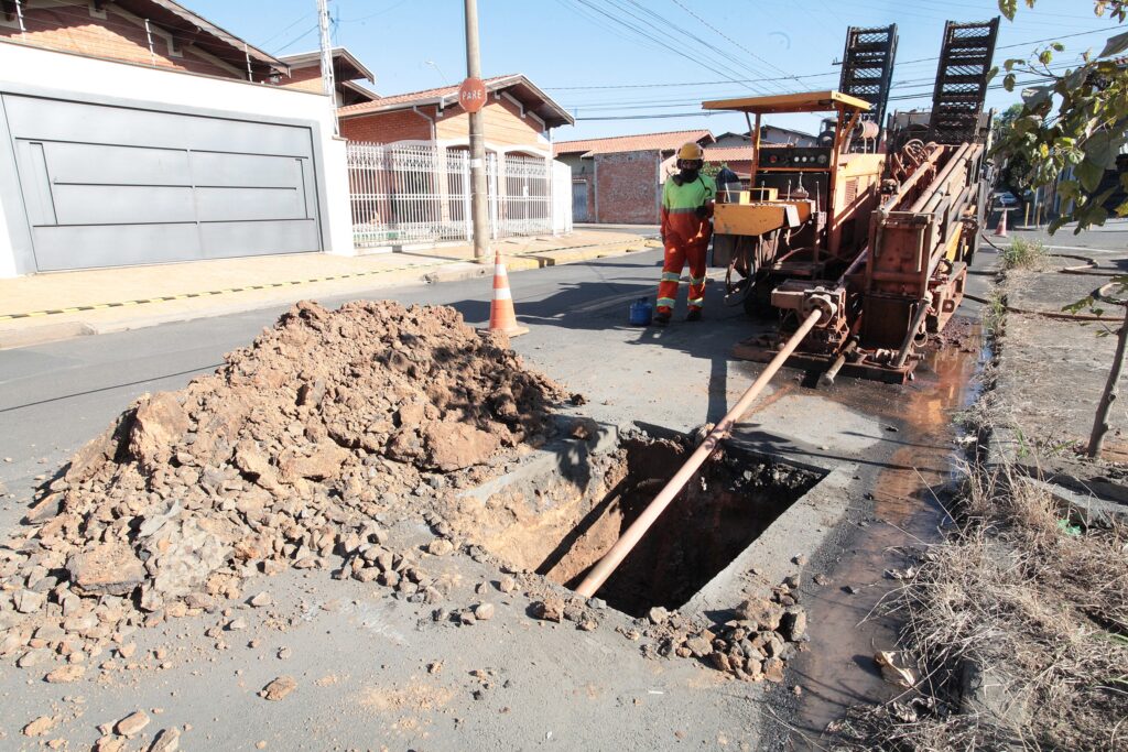 Uma cena de canteiro de obras em uma rua residencial mostra um trabalhador operando uma máquina de perfuração ao lado de uma vala aberta e um monte de terra sob um céu claro e ensolarado. A imagem apresenta um cenário de trabalho em uma rua residencial sob um céu azul claro e ensolarado. Em primeiro plano, no lado esquerdo, há um grande monte de terra e pedras de tonalidade marrom-avermelhada, indicando material escavado da rua. O solo parece seco e compactado em algumas partes, com fragmentos de rocha espalhados. Ao lado deste monte de terra, no centro da imagem, uma vala retangular foi aberta no asfalto escuro, revelando o subsolo mais escuro e úmido. Um cano de cor clara, possivelmente PVC, estende-se diagonalmente para baixo dentro da vala. Ao redor da vala e na entrada do cano, há acúmulo de água barrenta. Na superfície da rua, à direita do monte de terra, um cone de trânsito laranja e branco serve como sinalização. O asfalto da rua é de cor cinza escuro, com algumas manchas de sujeira e pequenas pedras espalhadas pela atividade de construção. Mais à direita, um trabalhador vestido com uniforme de segurança está de pé ao lado de uma máquina de perfuração grande e robusta. O trabalhador usa um macacão laranja com faixas refletivas, uma camisa de mangas compridas verde-limão por baixo, um capacete de segurança amarelo e um cachecol ou máscara escura cobrindo a parte inferior do rosto. Suas botas são escuras. A máquina, de cor amarela e laranja-ferrugem, tem diversas peças mecânicas expostas, braços e o que parecem ser duas estruturas elevadas e perfuradas na parte traseira, provavelmente para guiar o equipamento de perfuração. Há poças de água barrenta e lama ao redor da base da máquina e na rua, refletindo fragmentos do céu claro. Um pequeno recipiente azul está no chão, perto dos pés do trabalhador. Atrás da área de trabalho, no lado esquerdo da rua, há uma residência com um grande portão de garagem metálico de cor cinza claro, com um design moderno de painéis horizontais e verticais, em uma parede branca. Acima do portão, observa-se uma parede de tijolos e um telhado de telhas cerâmicas avermelhadas, com fios de segurança no topo. À direita desta casa, outra casa de tijolos com telhado de telhas e uma cerca branca de ferro forjado em estilo ornamental se destacam. Um poste de utilidade pública de concreto está erguido em frente a esta casa, exibindo uma placa octogonal de "PARE" em tom marrom. Fios elétricos e de comunicação cruzam o céu azul, criando linhas diagonais e horizontais na parte superior da imagem. A calçada no lado direito da rua possui grama seca e algumas plantas ralas. Um pequeno córrego de água barrenta flui ao longo da sarjeta, refletindo a luz do sol. Uma árvore com folhas verdes escuras pode ser vista no canto superior direito, obscurecendo ligeiramente a vista de uma parte da rua de trás. A luz solar é intensa, com sombras nítidas projetadas pela máquina, pelo trabalhador, pelo poste e pelos edifícios, indicando um dia claro e sem nuvens. A atmosfera geral é de trabalho em andamento, com a funcionalidade da obra contrastando com o ambiente residencial ao redor.