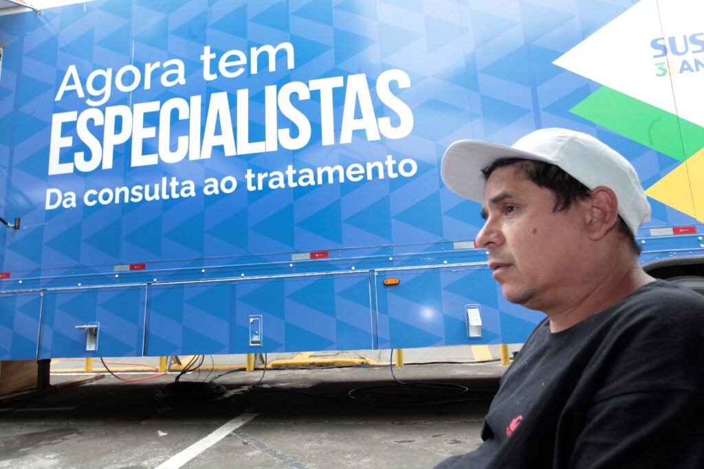 A imagem mostra um homem de perfil, usando um boné branco e uma camiseta escura, olhando para o lado. Atrás dele, há um veículo grande, possivelmente uma unidade móvel de saúde, pintado em tons de azul com um design geométrico. No lado do veículo está escrito, em letras grandes e brancas: "Agora tem ESPECIALISTAS" e "Da consulta ao tratamento". Também aparece o logo do SUS com a indicação "35 ANOS". A cena parece estar ao ar livre, em um local com piso asfaltado, possivelmente um estacionamento ou rua. A imagem transmite a ideia de um serviço de saúde móvel que oferece atendimento especializado, destacando a presença do SUS.