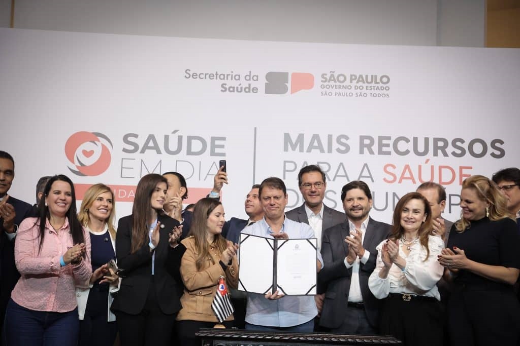 A imagem mostra um grupo de pessoas, provavelmente autoridades ou representantes do governo, reunidas em um palco para uma cerimônia oficial. No centro da foto, um homem veste uma camisa azul clara e segura um documento aberto em uma pasta preta, sorrindo para a câmera. Ao seu redor, outras pessoas estão alinhadas, algumas aplaudindo, todas vestidas de forma formal, com ternos, camisas sociais e blusas. Ao fundo, há uma parede branca com os logotipos e textos oficiais do governo do Estado de São Paulo, incluindo "Secretaria da Saúde", "Governo do Estado de São Paulo", "Saúde em Dia" e "Mais Recursos para a Saúde". A iluminação é clara e uniforme, típica de eventos oficiais realizados em ambientes fechados. A imagem transmite um momento de anúncio ou assinatura importante relacionado à área da saúde pública, evidenciando uma ação governamental em prol do setor.
