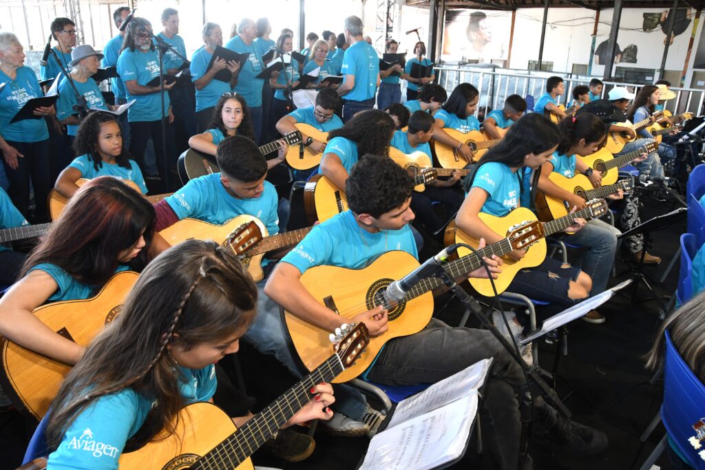 A imagem mostra um grande grupo de pessoas, principalmente jovens estudantes, participando de uma oficina ou ensaio musical em um ambiente interno amplo. A maioria dos estudantes está sentada em fileiras, tocando violões acústicos e usando camisetas azuis vibrantes com a palavra "Aviagen" estampada. Eles parecem concentrados, muitos olhando para partituras apoiadas em suportes à sua frente. Atrás deles, há um grupo de adultos em pé, segurando pastas ou partituras, possivelmente integrantes de um coro ou da equipe organizadora. O local parece ser um salão grande com teto alto e estrutura metálica aparente, com iluminação clara e uniforme. Também há microfones posicionados na frente dos estudantes, indicando que pode estar ocorrendo uma apresentação ou gravação. A imagem transmite uma atmosfera de aprendizado coletivo e dedicação à música.