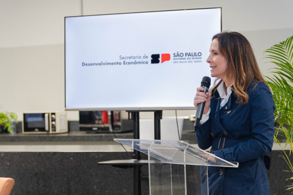 A imagem mostra uma mulher falando em um microfone, posicionada atrás de um púlpito transparente. Ela veste um casaco azul escuro com cinto e uma camisa branca por baixo. Ao fundo, há uma tela digital com o logotipo e o nome da "Secretaria de Desenvolvimento Econômico" do governo do estado de São Paulo. O ambiente parece ser interno, possivelmente uma sala de eventos ou uma área de escritório, com uma planta verde à direita e um balcão com alguns objetos, como um micro-ondas, ao fundo. A mulher aparenta estar fazendo uma apresentação ou discurso formal em um evento oficial.