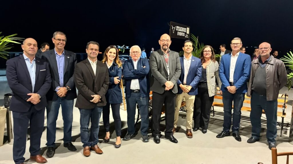 A imagem mostra um grupo de dez pessoas alinhadas em um ambiente externo à noite. São oito homens e duas mulheres, todos vestidos de forma elegante, com roupas que variam entre blazers, camisas sociais, suéteres e calças. Eles estão posando para a foto, a maioria com sorrisos discretos. Ao fundo, há uma placa iluminada com o texto "STEAKHOUSE EVENTOS", indicando que provavelmente estão em um local destinado a eventos ou celebrações. O cenário é um espaço aberto, possivelmente um pátio, com iluminação artificial que destaca bem as pessoas, enquanto o fundo permanece mais escuro, com algumas plantas visíveis nas laterais. A atmosfera sugere um encontro formal ou uma celebração profissional.