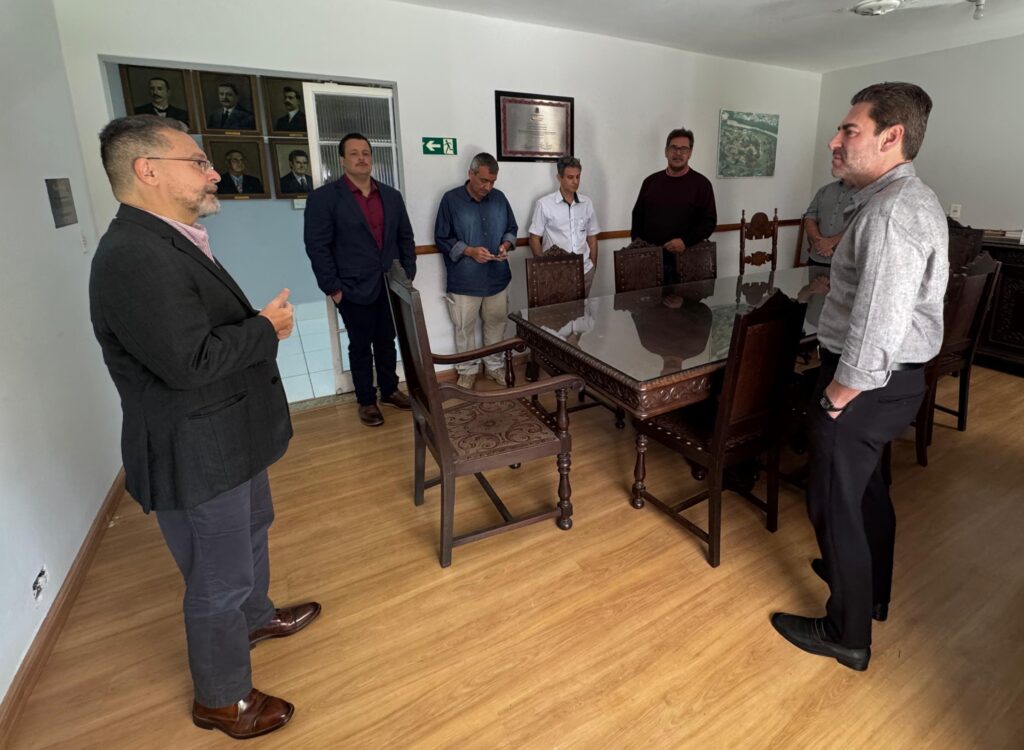 A imagem mostra um grupo de seis homens reunidos em uma sala de reuniões com decoração sóbria. * No centro da sala, há uma mesa retangular de madeira escura com tampo de vidro, cercada por cadeiras de estilo clássico com assentos estofados. * Dois homens estão em destaque nas extremidades da mesa: um à esquerda, vestindo paletó escuro e óculos, gesticulando como se estivesse conduzindo uma apresentação ou conversa; e outro à direita, de camisa cinza e calças pretas, observando atentamente. * Ao fundo, junto à parede, quatro outros homens permanecem em pé, alguns observando a interação e outros olhando para baixo. * A decoração da sala inclui uma série de retratos emoldurados na parede esquerda, uma placa comemorativa na parede central, uma placa de saída de emergência e um quadro decorativo com uma imagem paisagística. O piso é de madeira clara, conferindo um tom formal ao ambiente.