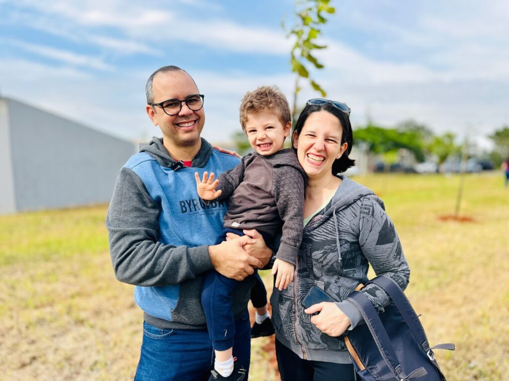 Nesta foto, uma família de três pessoas sorri alegremente em um campo aberto em um dia ensolarado. • À esquerda, o pai, usando óculos, um moletom azul e cinza da marca "Byron Bay" e calça jeans, segura o filho no colo. • No centro, o menino, de cabelos cacheados, veste um casaco marrom e calça azul, e acena para a câmera com um sorriso largo. • À direita, a mãe, de cabelos curtos, usa um casaco cinza estampado com capuz e carrega uma mochila preta na mão. O fundo mostra uma área gramada ampla sob um céu azul com algumas nuvens, sugerindo um parque ou um local de atividades ao ar livre.
