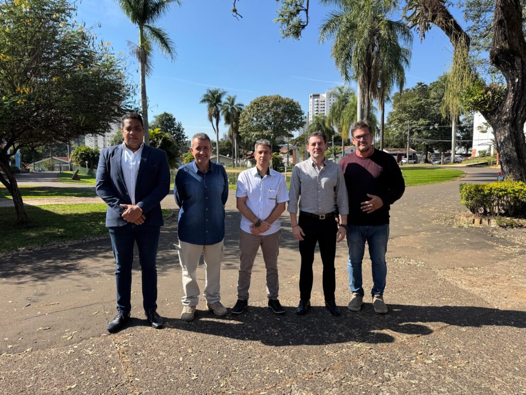 A imagem apresenta cinco homens posando para uma foto ao ar livre, em um ambiente que parece ser um parque ou área de lazer com gramado, árvores e calçamento de terra batida. * Da esquerda para a direita, o primeiro homem veste um blazer azul-marinho, camisa social branca, calça jeans escura e sapatos sociais pretos. * O segundo homem usa uma camisa de botão em tom de azul escuro, calça clara e tênis. * O terceiro homem, posicionado no centro, veste uma camisa polo branca com detalhes na gola, calça bege e tênis escuros. * O quarto homem está de camisa social cinza, calça preta com cinto e sapatos sociais pretos. * O quinto homem, à direita, veste um suéter de gola careca em tom bordô, calça jeans azul e tênis claros. O cenário é iluminado pela luz do dia, com sombras projetadas no chão, sugerindo um período de sol forte. Ao fundo, é possível observar vegetação, palmeiras e algumas construções ao longe.
