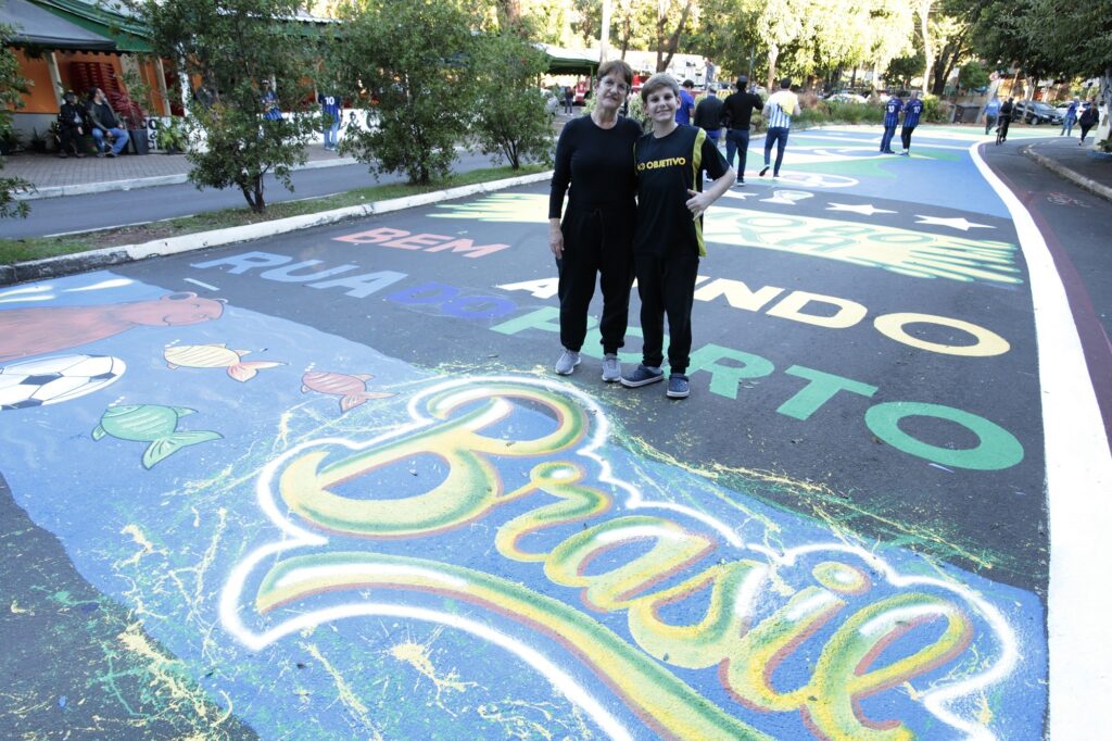 A imagem mostra uma cena ao ar livre, provavelmente numa rua ou praça pública, onde o chão foi pintado com um mural colorido e vibrante. No centro da imagem, há uma mulher adulta e um menino, ambos de cabelos escuros, posando lado a lado para a foto. A mulher veste roupa preta, enquanto o menino usa uma camiseta preta com a palavra "OBJETIVO" escrita em amarelo. O chão está decorado com letras grandes e coloridas, em tons vivos como amarelo, verde, azul e vermelho. Entre as palavras pintadas no asfalto, destacam-se "BEM", "RUA", "MUNDO", "PORTO" e "Brasil", esta última em destaque com as cores da bandeira brasileira (amarelo, verde e azul). Também há ilustrações, como uma bola de futebol e peixes, remetendo possivelmente a temas culturais ou esportivos do Brasil. Ao fundo, há árvores, um prédio com uma cobertura laranja e algumas pessoas caminhando ou paradas, indicando que o local pode estar sendo usado para um evento comunitário ou celebração. A luz do dia é forte, evidenciando um dia ensolarado. A imagem transmite uma atmosfera alegre e festiva, valorizando a cultura brasileira e o espaço público.