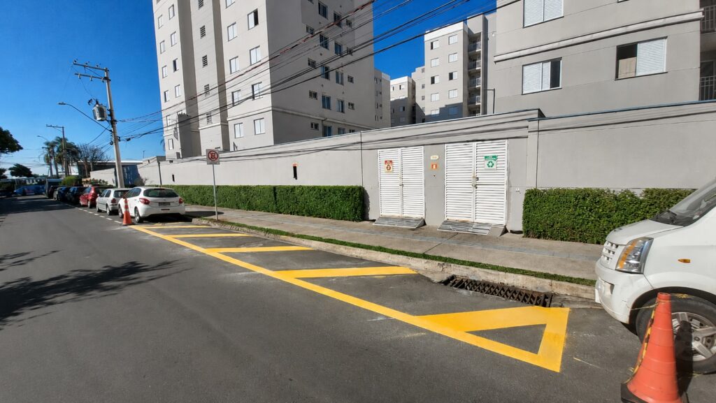Esta foto mostra uma rua lateral de um condomínio residencial em um dia ensolarado. Sinalização: O destaque é uma área pintada no asfalto com faixas amarelas diagonais, indicando uma zona de proibição de estacionamento ou área de manobra. Há um cone de trânsito laranja posicionado logo à frente de um carro branco estacionado à direita. O condomínio: Ao fundo, vemos grandes edifícios residenciais de cor cinza. O muro que cerca o condomínio é longo, também cinza, com sebes verdes bem aparadas logo abaixo. Estrutura: Há dois portões metálicos brancos inseridos no muro e uma placa de "proibido estacionar" visível na calçada. A rua parece ser residencial e tranquila, com outros carros estacionados ao longo da via à esquerda.