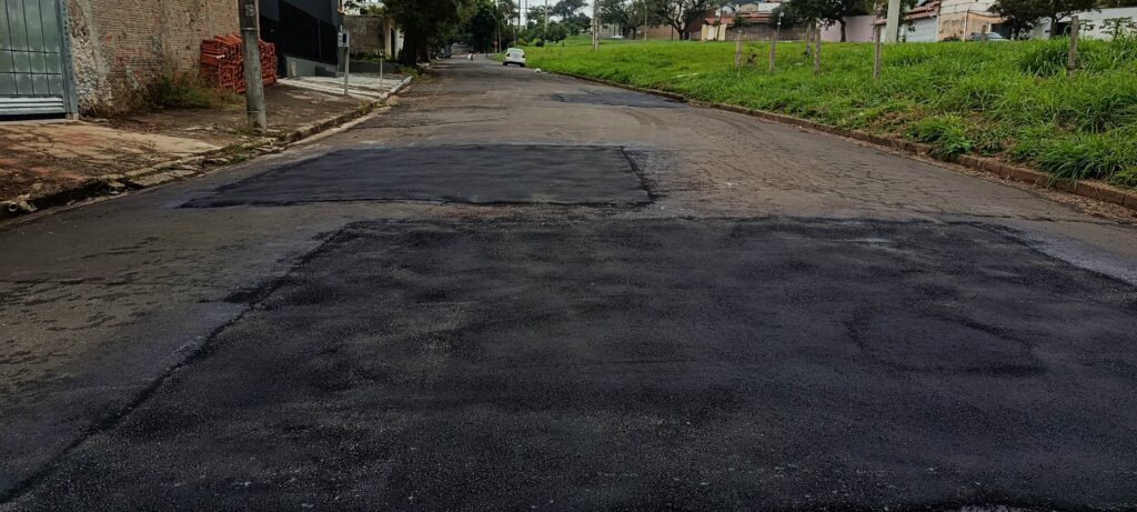 Esta é uma foto de uma rua residencial que parece ter passado por reparos recentes no asfalto. Em primeiro plano, é possível ver grandes áreas de asfalto novo, bem preto e uniforme, cobrindo trechos onde provavelmente havia buracos ou valas. O restante da rua mostra um pavimento mais antigo, com desgaste e algumas rachaduras visíveis. Nas laterais, há uma calçada simples à esquerda com materiais de construção acumulados e, à direita, um gramado verde que segue ao longo da rua. Ao fundo, a via se estende em direção a algumas casas e parece haver um carro trafegando à distância. O céu está nublado, dando um tom cinzento à cena.