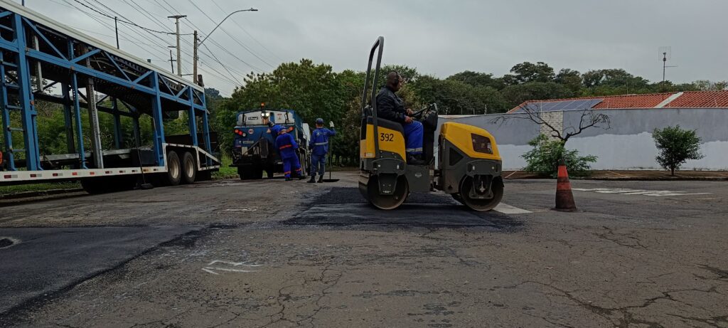 Nesta cena, vemos uma equipe trabalhando no recapeamento ou reparo de uma via. Em destaque, um rolo compactador amarelo (com o número 392) está sendo operado por um trabalhador, passando sobre um trecho de asfalto recém-aplicado, que aparece bem escuro e liso. Ao fundo, há um caminhão de serviço e outros dois trabalhadores uniformizados, provavelmente ajustando o trabalho na pista. À esquerda, vê-se a lateral de uma carreta cegonha estacionada. O local parece ser uma área externa com árvores e um muro ao fundo sob um céu nublado, indicando um dia típico de trabalho de manutenção urbana.