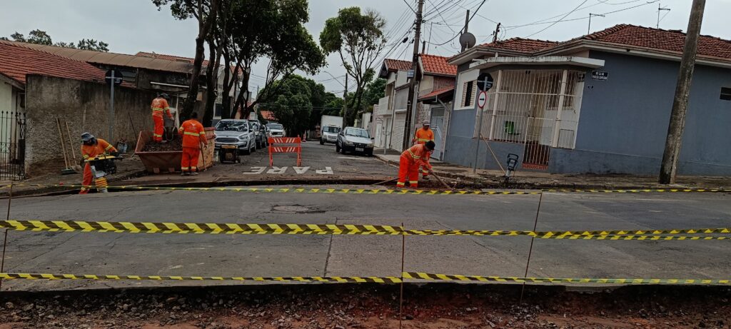 A imagem mostra uma cena de obras em uma rua residencial. Quatro trabalhadores, vestidos com uniformes laranja fluorescentes, estão realizando serviços na via. Um deles está operando uma compactadora de solo, enquanto os outros trabalham com ferramentas manuais. Há um grande contêiner metálico para descarte de materiais próximo aos trabalhadores. A rua está parcialmente escavada e isolada por uma fita de advertência amarela e preta. Ao fundo, é possível ver carros estacionados e casas com telhados de cerâmica. No asfalto está pintada a palavra "PARE" e uma placa indica o nome da rua: "Rua Princesa Leopoldina". O céu está nublado, indicando um dia encoberto. A cena transmite uma atmosfera típica de manutenção urbana em andamento.