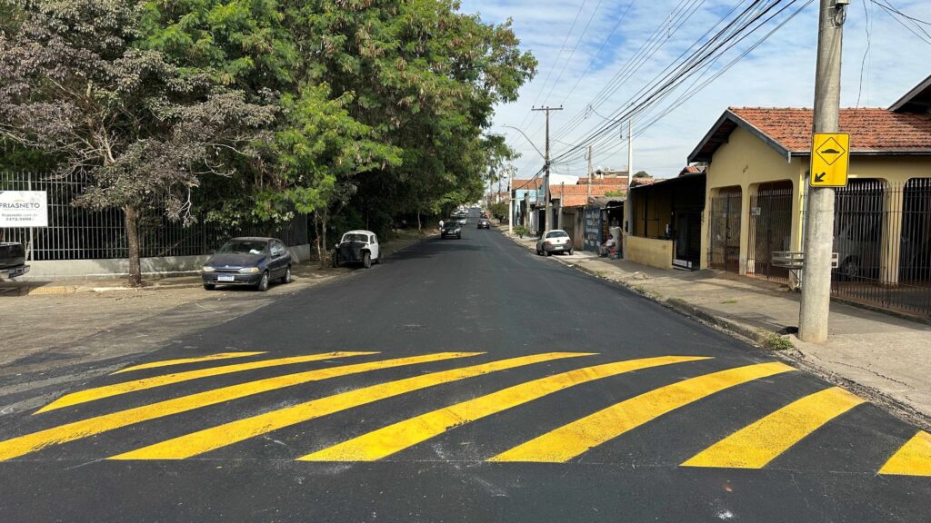 A imagem mostra uma rua residencial pavimentada recentemente, com um quebra-molas pintado em amarelo vibrante no centro da via. À direita, há um poste de concreto com uma placa amarela de advertência indicando a presença do quebra-molas. De um lado da rua, há várias casas com telhados de cerâmica vermelha e janelas protegidas por grades metálicas. Do outro lado, uma área com árvores verdes e carros estacionados, incluindo um Fusca branco antigo e um carro hatch azul escuro. O céu está parcialmente nublado, com boa iluminação natural, sugerindo um dia ensolarado. Também é possível ver uma placa de imobiliária com o nome "FRIASNETO" e um site e telefone de contato. A perspectiva da foto é centralizada na rua, levando o olhar para o horizonte onde há mais veículos.