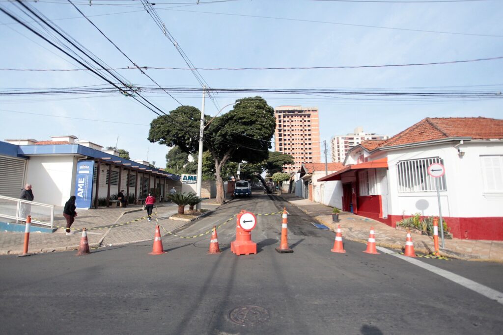 A imagem mostra uma rua urbana durante o dia, parcialmente bloqueada por cones de trânsito laranja e uma fita amarela de advertência, indicando que o acesso está restrito ou em manutenção. No centro da rua, há uma grande árvore que chama atenção, cercada pelos cones que orientam os veículos a manterem-se à esquerda. À esquerda da imagem, há um edifício com uma placa que diz "AME - Ambulatório Médico de Especialidades", sugerindo que é uma unidade de saúde. Algumas pessoas estão próximas à entrada desse prédio. À direita, há uma construção residencial com janelas e varandas, pintada em tons claros com detalhes vermelhos. Ao fundo, é possível ver prédios maiores, possivelmente apartamentos, alguns deles ainda em construção ou reforma. O céu está limpo e azul, e a iluminação natural destaca todos os elementos da cena. Fios elétricos cruzam a parte superior da imagem, comuns em áreas urbanas. A rua parece estar em uma área movimentada, mas o bloqueio indica algum tipo de intervenção temporária.