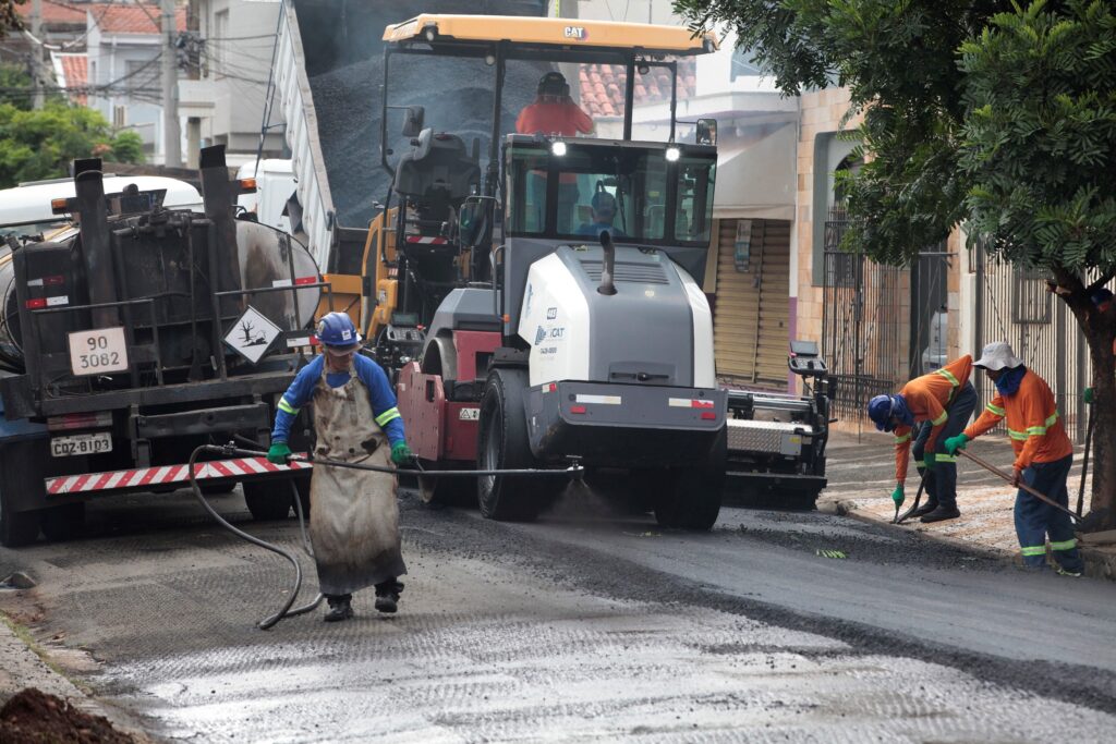 A imagem mostra uma cena de construção de estrada em uma área residencial. No centro, há uma máquina de pavimentação de asfalto da marca CAT, que está aplicando uma nova camada de asfalto preto na rua. À esquerda, há um caminhão tanque grande, com os números "90 3082" e a placa "CQZ-8103", provavelmente usado para transportar materiais para a pavimentação. No primeiro plano, um trabalhador usa um capacete azul e um avental de couro pesado para aplicar um líquido na superfície da estrada com uma ferramenta longa, possivelmente um spray para preparar ou tratar o asfalto. À direita, outros dois trabalhadores com coletes laranja fluorescentes e capacetes azuis estão usando ferramentas manuais para alisar ou limpar as bordas do asfalto recém-aplicado. O ambiente é urbano ou suburbano, com árvores e edifícios ao fundo, e o dia parece estar nublado, com luz difusa. A imagem transmite uma sensação de trabalho em equipe e esforço coordenado para realizar a pavimentação da rua.