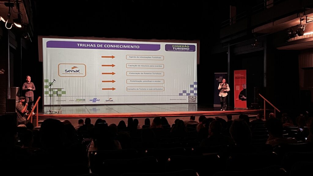 Nesta imagem, vemos um auditório escuro durante uma apresentação. No palco, há um telão grande que exibe o tema "Trilhas de Conhecimento" do evento "Conexão Turismo". O telão lista cinco tópicos principais, acompanhados por setas laranjas: • Agente de Informações Turísticas • Captação de recursos para eventos • Elaboração de Roteiros Turísticos • Roteirização: precificar e vender • Conselho de Turismo e suas atribuições No canto esquerdo do telão, aparece a logo do Senac Atendimento Corporativo. Na parte inferior, há os logos da Prefeitura de Piraí, Sebrae e Senac. Sobre o palco, vemos: • À esquerda, um homem de pé perto de um suporte de microfone, possivelmente um dos organizadores ou um palestrante. • À direita, uma mulher de pé, segurando um microfone, apresentando o conteúdo. • Há banners nas laterais do palco, um azul (Sebrae) e outro vermelho (Soluções Educacionais). • O público está sentado no auditório, assistindo à palestra.