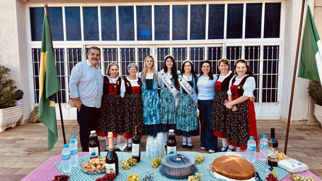 Esta imagem mostra um grupo de 10 pessoas posando para uma foto ao ar livre, em frente a uma estrutura com grandes janelas de vidro e grades de metal. O cenário é festivo, com uma mesa em primeiro plano coberta por uma toalha xadrez verde e branca, contendo garrafas de vinho, suco, copos descartáveis, uvas, petiscos e um bolo. * À esquerda, um homem de camisa social clara e calça escura está ao lado de duas senhoras vestidas com trajes típicos tradicionais, que incluem coletes escuros sobre blusas brancas e saias estampadas. * No centro, três jovens mulheres também usam trajes típicos similares, sendo que duas delas exibem faixas com os títulos de "Rainha" e "Princesa", indicando uma celebração cultural ou comunitária. * À direita, completando o grupo, há uma mulher vestindo roupas casuais (blusa branca e calça jeans) e outras duas senhoras também trajadas com as vestimentas tradicionais, semelhantes às das primeiras senhoras.
