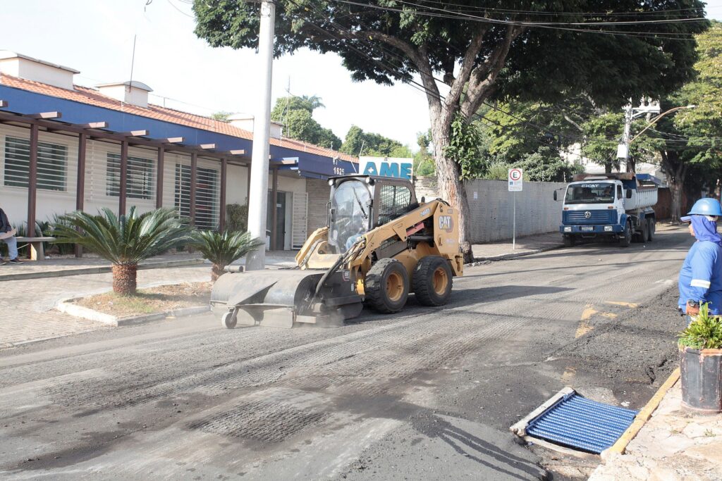 A imagem mostra uma cena de trabalho de manutenção viária durante o dia. No centro, há uma máquina amarela da marca Caterpillar, do tipo carregadeira compacta, equipada com um acessório para fresagem do asfalto. A máquina está em operação, removendo a camada superior da pavimentação da rua, o que gera uma nuvem de poeira. À direita, um trabalhador vestido com uniforme azul, colete refletivo e capacete azul observa a atividade. Ao fundo, há um caminhão basculante branco e azul estacionado próximo a um muro. Do lado esquerdo da imagem, vê-se uma construção com fachada azul e algumas palmeiras pequenas em um canteiro. Também há uma placa de sinalização de estacionamento e postes com fiação elétrica visível no céu claro. A cena transmite a ideia de um trabalho urbano de reparo ou renovação da via pública.