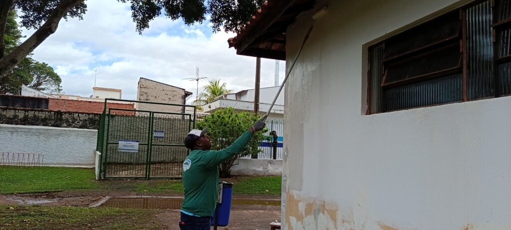 A imagem mostra um trabalhador vestido com uniforme verde e boné branco pintando a parede externa de um prédio. Ele está usando um rolo de pintura com cabo longo para alcançar uma área alta da parede, próxima ao beiral do telhado. A parede é branca, mas apresenta algumas áreas desgastadas e manchas perto da base. Ao lado da parede, há uma janela com persianas marrons. No fundo, é possível ver um portão verde, uma lixeira azul e algumas árvores. O céu está nublado, indicando um dia com pouca luz solar direta. Na parede, há um letreiro escrito "ESTAÇÃO ELEVATÓRIA DE ESGOTO MORUMBI", indicando que o local é uma estação de bombeamento de esgoto. A cena transmite uma atividade de manutenção ou reforma do local.