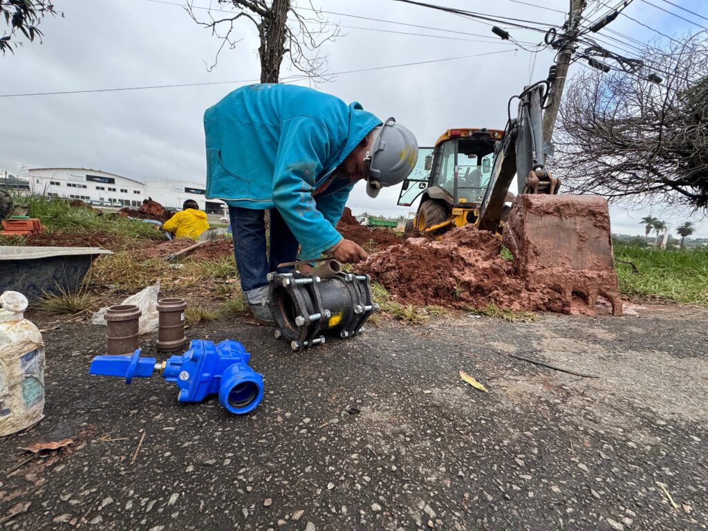 A imagem mostra uma cena de construção ao ar livre em um dia nublado. No centro da imagem, há um trabalhador vestido com uma jaqueta azul brilhante com capuz, calças jeans escuras e um capacete cinza, agachado e manuseando uma peça metálica de tubulação no chão asfaltado. Ao lado dele, há uma válvula azul e dois segmentos de tubos marrons. Ao fundo, um trator amarelo da marca John Deere está próximo a uma pilha de terra avermelhada escavada. Outro trabalhador, vestido com uma jaqueta amarela de alta visibilidade, aparece mais ao fundo, próximo a uma vala aberta. Ao fundo, há um prédio industrial grande com a inscrição "LADO A LADO" e algumas árvores. O céu está nublado, indicando um dia sem sol direto. A imagem transmite uma atividade de manutenção ou instalação de infraestrutura, possivelmente relacionada a tubulações subterrâneas.