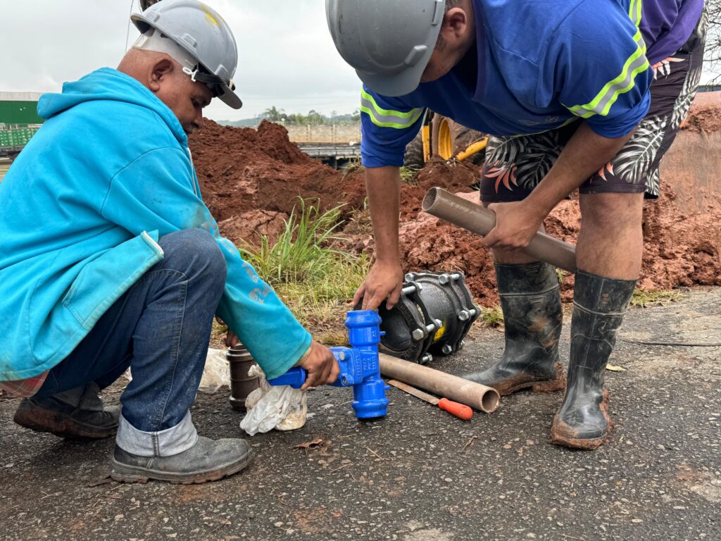 A imagem mostra dois trabalhadores da construção civil em um canteiro de obras ao ar livre. O trabalhador à esquerda está agachado, vestindo um moletom azul, calça jeans, capacete branco e botas de trabalho escuras. Ele segura uma peça azul, provavelmente um componente de encanamento. O trabalhador à direita está em pé, levemente inclinado para frente, usando uma camiseta azul com faixas refletivas, shorts estampados, capacete cinza e botas pretas de borracha cobertas de barro. Ele segura um tubo de PVC marrom, próximo a uma grande abraçadeira metálica preta no chão de asfalto. Ao redor deles, há terra avermelhada e vegetação esparsa, sugerindo que estão instalando ou reparando uma tubulação. O ambiente está iluminado por luz natural difusa, típica de um dia nublado.