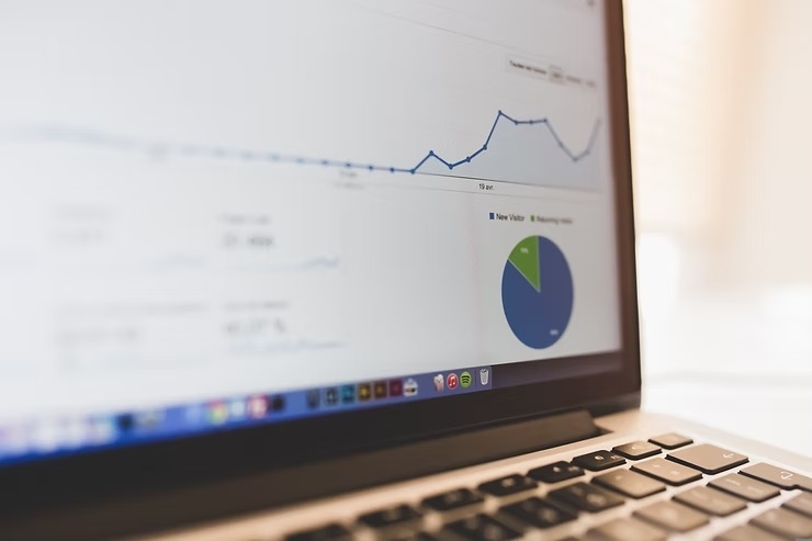 A imagem mostra um close-up da tela de um laptop, onde está aberto um painel do Google Analytics. Na tela, é possível ver um gráfico de linhas que indica uma tendência de crescimento e um gráfico de pizza que apresenta dados sobre visitantes, com as categorias "New Visitor" (Novo Visitante) e "Returning Visitor" (Visitante Retornando). A imagem está focada principalmente na tela do laptop, enquanto o teclado em primeiro plano está desfocado. A iluminação é suave e natural, sugerindo um ambiente de trabalho, possivelmente em casa ou escritório. O laptop parece ser um MacBook, identificado pela barra de aplicativos na parte inferior da tela. A composição da foto, com o ângulo lateral e o foco seletivo, destaca o conteúdo da tela e cria uma sensação de profundidade.