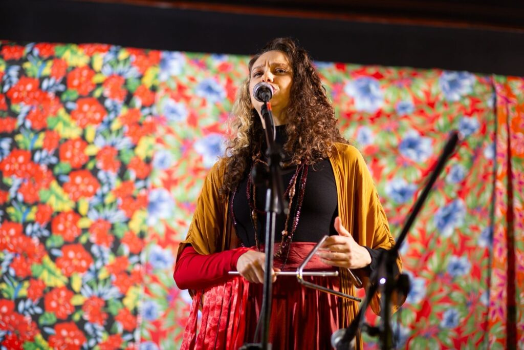 A imagem retrata uma musicista em um palco, capturada em um plano médio, enquanto canta e toca um triângulo. * Ela possui cabelos longos, cacheados e volumosos, com mechas claras, e veste uma blusa preta de gola alta sob um quimono ou capa em tom de mostarda. Complementando o visual, utiliza um colar longo com detalhes em madeira ou contas, e uma saia avermelhada. * A artista está posicionada centralmente, cantando em um microfone montado em um pedestal. Com a mão direita, ela segura o instrumento de percussão (triângulo), enquanto a esquerda parece estar em movimento, acompanhando o ritmo. * O fundo é composto por um painel ou cortina vibrante, estampado com um padrão floral denso e colorido, predominando flores vermelhas e azuis sobre um fundo escuro, o que confere um aspecto cultural ou folclórico à cena. A iluminação é focada na artista, criando um contraste suave com o fundo colorido.