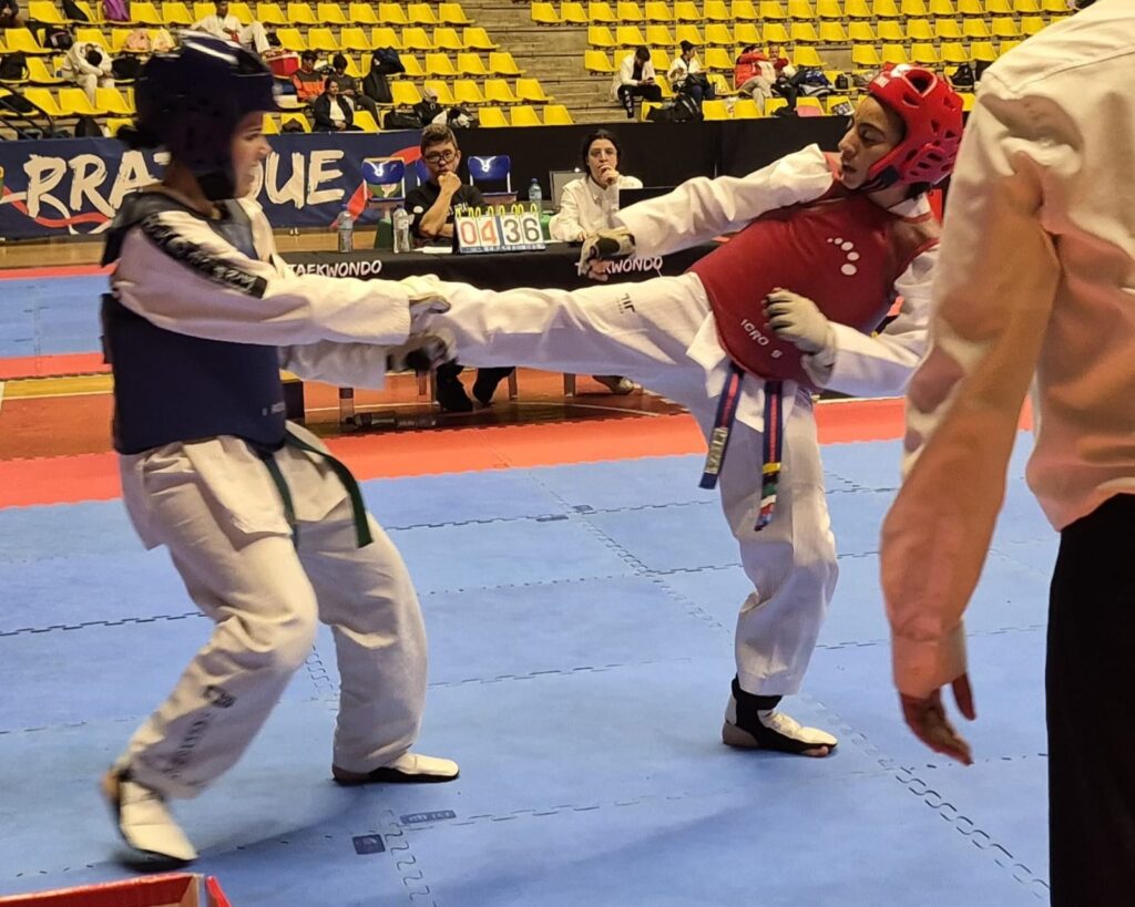 A imagem mostra uma cena de uma luta de taekwondo em um ambiente esportivo indoor. Duas atletas femininas estão em ação, ambas vestindo doboks brancos e equipamentos de proteção, incluindo capacetes e coletes protetores — uma com colete azul e capacete azul, a outra com colete vermelho e capacete vermelho. A atleta de vermelho está executando um chute alto em direção à adversária de azul. Um árbitro, vestido com camisa branca e calça preta, observa atentamente a luta. Ao fundo, há uma mesa com juízes e uma placa eletrônica que exibe o placar e o tempo da luta. As arquibancadas amarelas estão vazias, indicando que o local não está cheio de espectadores naquele momento. O chão é revestido com tatames azuis, próprios para a prática do taekwondo, e há banners e inscrições relacionadas ao evento. A imagem transmite a intensidade e a concentração típicas de uma competição de artes marciais.