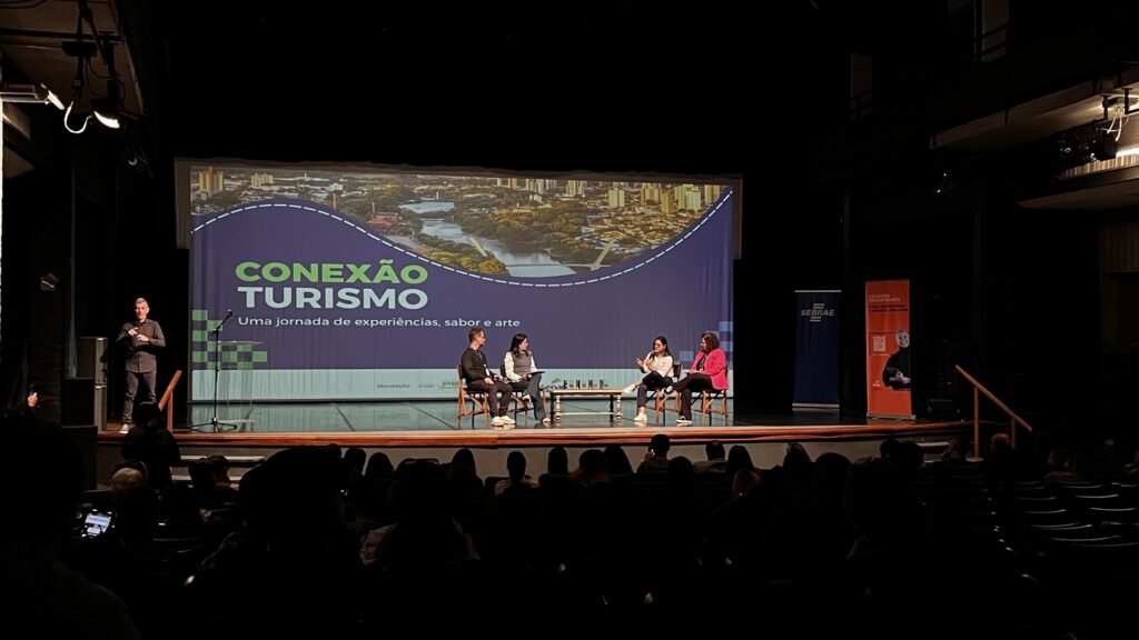 Nesta imagem, vemos um painel de debate ocorrendo em um auditório durante o evento "Conexão Turismo". No palco: • À esquerda, um homem de preto está em pé, fazendo a tradução em Libras para o público. • Ao centro, quatro pessoas estão sentadas em cadeiras, formando uma roda de conversa, com uma pequena mesa de centro entre elas. Elas parecem estar debatendo ou conversando com a plateia. • O telão ao fundo exibe o logo "CONEXÃO TURISMO", com o subtítulo "Uma jornada de experiências, sabor e arte", além de uma bela foto aérea de uma cidade cortada por um rio. • Nas laterais do palco, há banners do Sebrae e do Senac. A iluminação é focada no palco, mantendo o ambiente do auditório e a plateia na penumbra, criando um clima de atenção voltada para os debatedores.