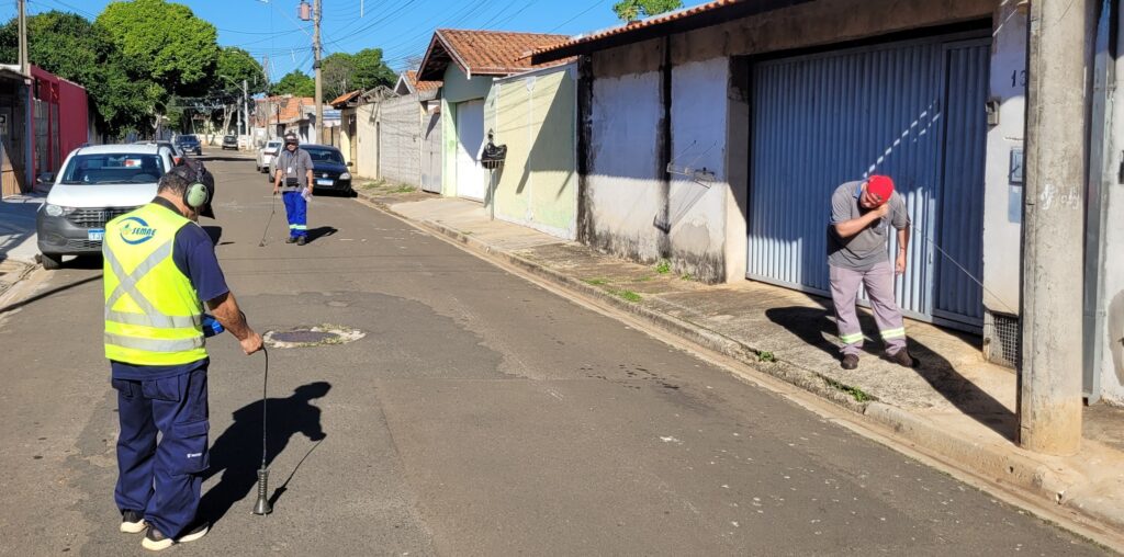 Esta é uma cena urbana em uma rua residencial onde dois trabalhadores estão realizando uma verificação ou inspeção técnica. Ação: O trabalhador em primeiro plano, à esquerda, veste um colete refletivo amarelo, camisa azul e calças cargo. Ele está segurando um dispositivo de detecção preso a uma haste, caminhando pela rua e monitorando algo no solo (parece ser a busca por vazamentos ou inspeção de tubulações subterrâneas). O outro trabalhador, mais ao fundo à esquerda, caminha na mesma direção com um equipamento semelhante. Detalhes Adicionais: Há um terceiro homem à direita, encostado em um poste e na parede de um portão de garagem, que parece estar verificando algo em um dispositivo ou cabo, ou talvez apenas fazendo uma pausa. Ambiente: A rua é asfaltada e parece ser uma zona residencial tranquila. Há casas com muros brancos, uma garagem de portão metálico cinza e alguns carros estacionados ao fundo. A iluminação é de um dia ensolarado, projetando sombras bem definidas no chão. Identificação: O colete do trabalhador em primeiro plano exibe o logotipo do "SEMAE" (Serviço Municipal de Água e Esgoto), indicando que se trata de uma equipe de manutenção ou fiscalização de serviços públicos de saneamento.